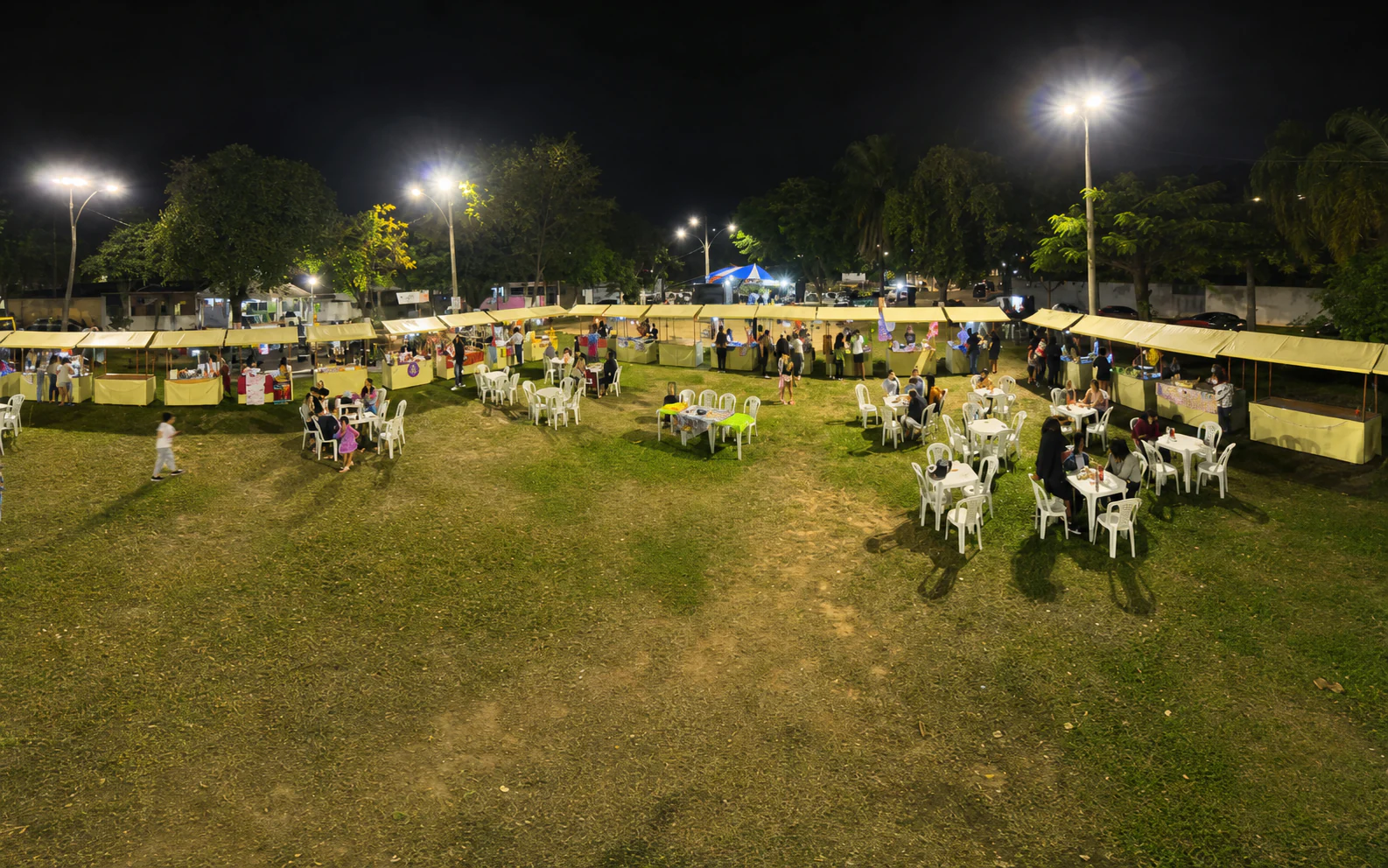 Vista noturna do Polo Gastronômico e Cultural João Petini, com barracas, mesas e visitantes na praça